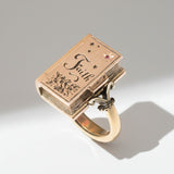 A rose gold ring with a sterling silver band and a ruby, featuring an intricate book design with the word 'Faith' and a star graphic.
