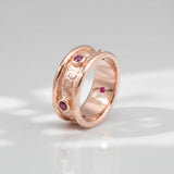 A rose gold band ring with hand engraved scrollwork and multiple small red gemstones set into the band.