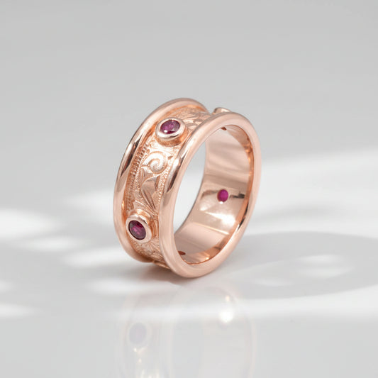 A rose gold band ring with hand engraved scrollwork and multiple small red gemstones set into the band.