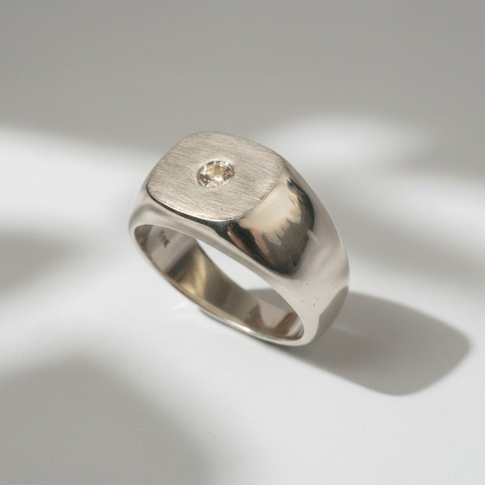 A solid 9-karat white gold signet-style ring with a matte emery finished face and a high polished body, set with a natural diamond.