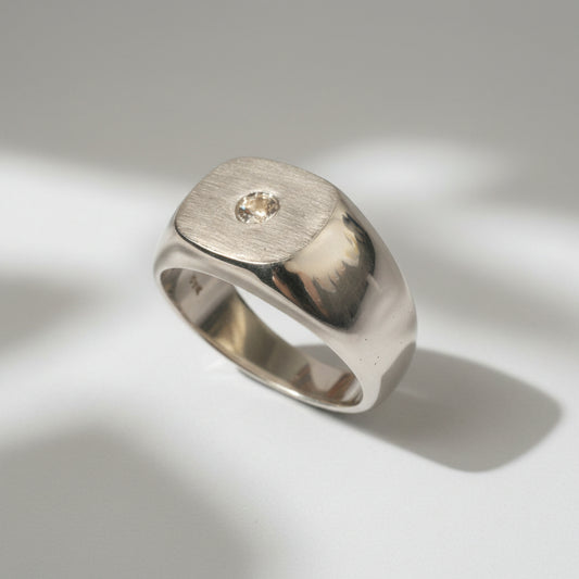 A solid 9-karat white gold signet-style ring with a matte emery finished face and a high polished body, set with a natural diamond.