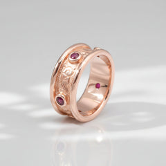 A rose gold band ring with hand engraved scrollwork and multiple small red gemstones set into the band.